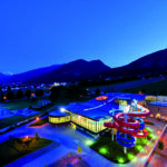 Luftaufnahme eines modernen Gebäudes mit einer roten Spiralrutsche, beleuchtet in der Abenddämmerung, vor der Kulisse von Bergen und einer Kleinstadt.