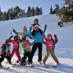 Een groep kinderen en volwassenen poseert samen op een besneeuwde skipiste, allemaal in skikleding, met een achtergrond van pijnbomen.