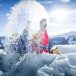 Twee mensen in winterkleding gooien speels sneeuw in de lucht op een besneeuwde berg met een helderblauwe lucht en bergachtig landschap op de achtergrond.