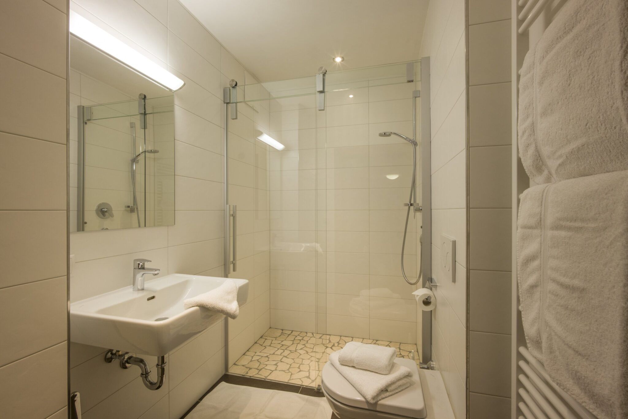 Modernes Badezimmer mit weiß gefliesten Wänden, ebenerdiger Dusche, wandmontiertem Waschbecken, Toilette und Handtuchhalter mit gefalteten Handtüchern.