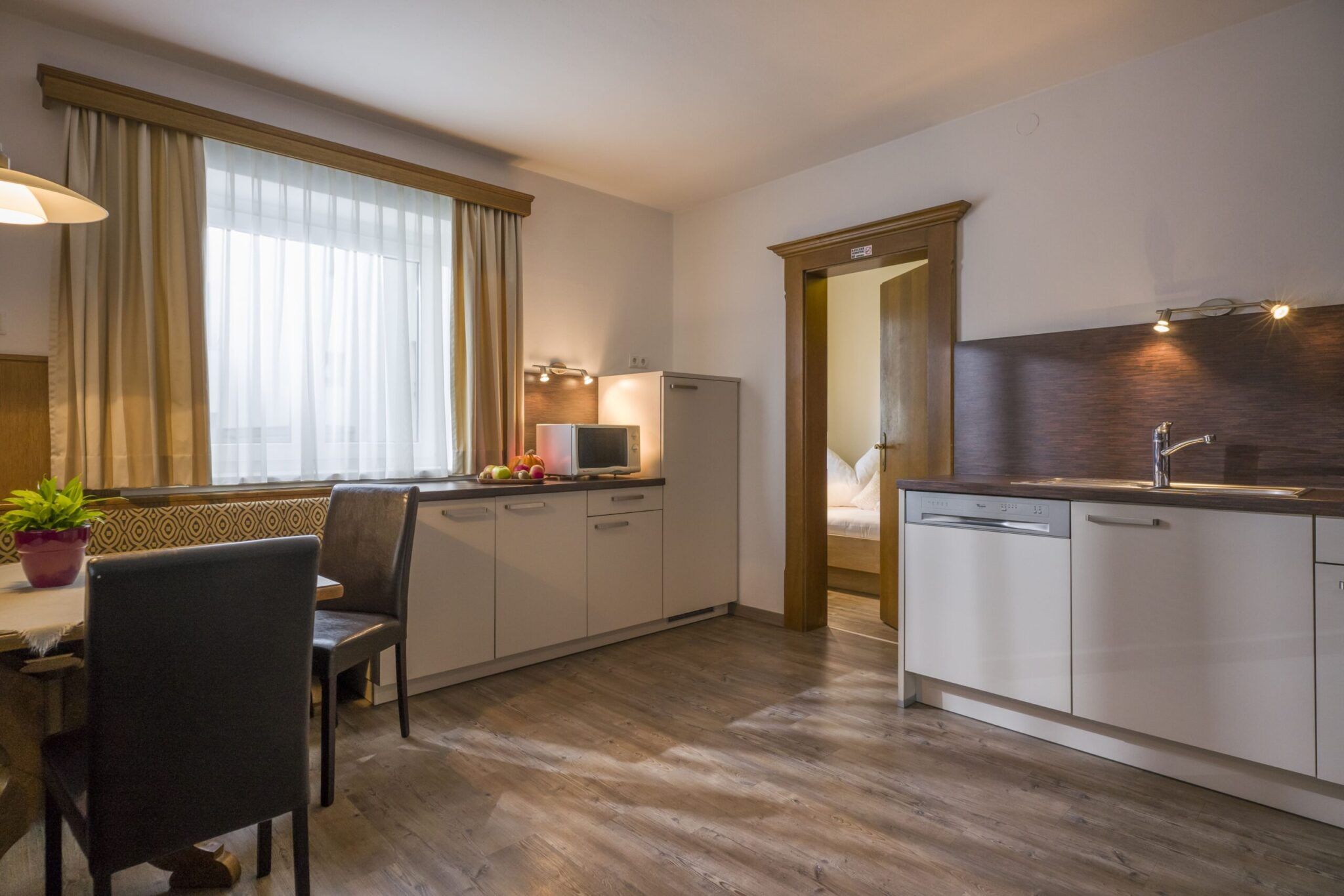 Moderne Küche mit Holzboden, weißen Schränken, Geräten aus Edelstahl, Esstisch und Durchgang zu einem Schlafzimmer.