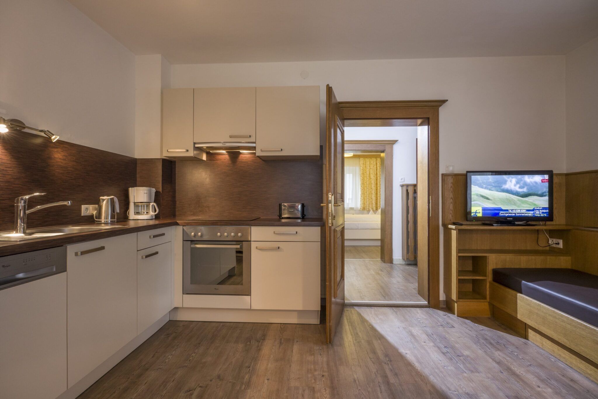 Eine moderne Küche mit weißen Schränken neben einem Wohnbereich mit Fernseher und Bank und einer offenen Tür zu einem Schlafzimmer.