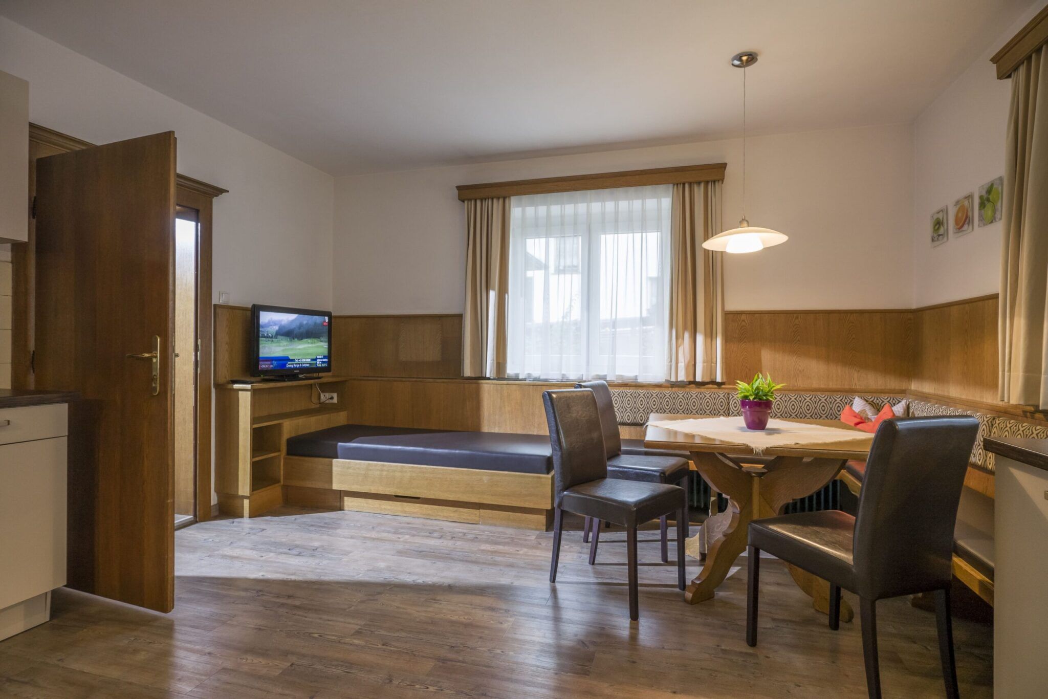Ein gemütlicher Essbereich mit Holztisch, Stühlen, Fernseher und einem Fenstersitz, beleuchtet von einer Hängelampe. Großes Fenster im Hintergrund.