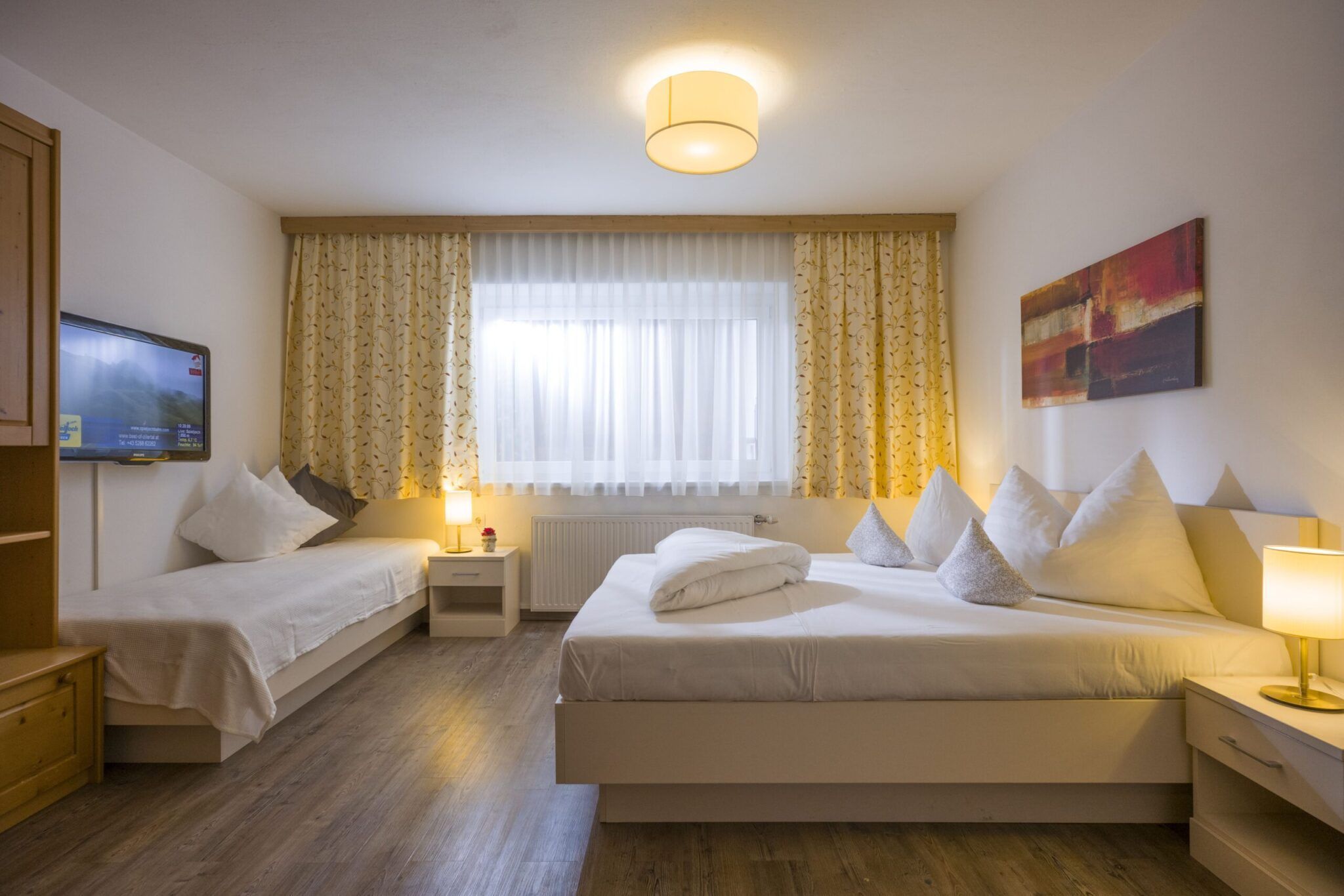 Ein gemütliches Schlafzimmer mit zwei Betten, einem Fernseher, Nachttischen, Lampen, gerahmten Kunstwerken und einem großen Fenster mit Vorhängen.