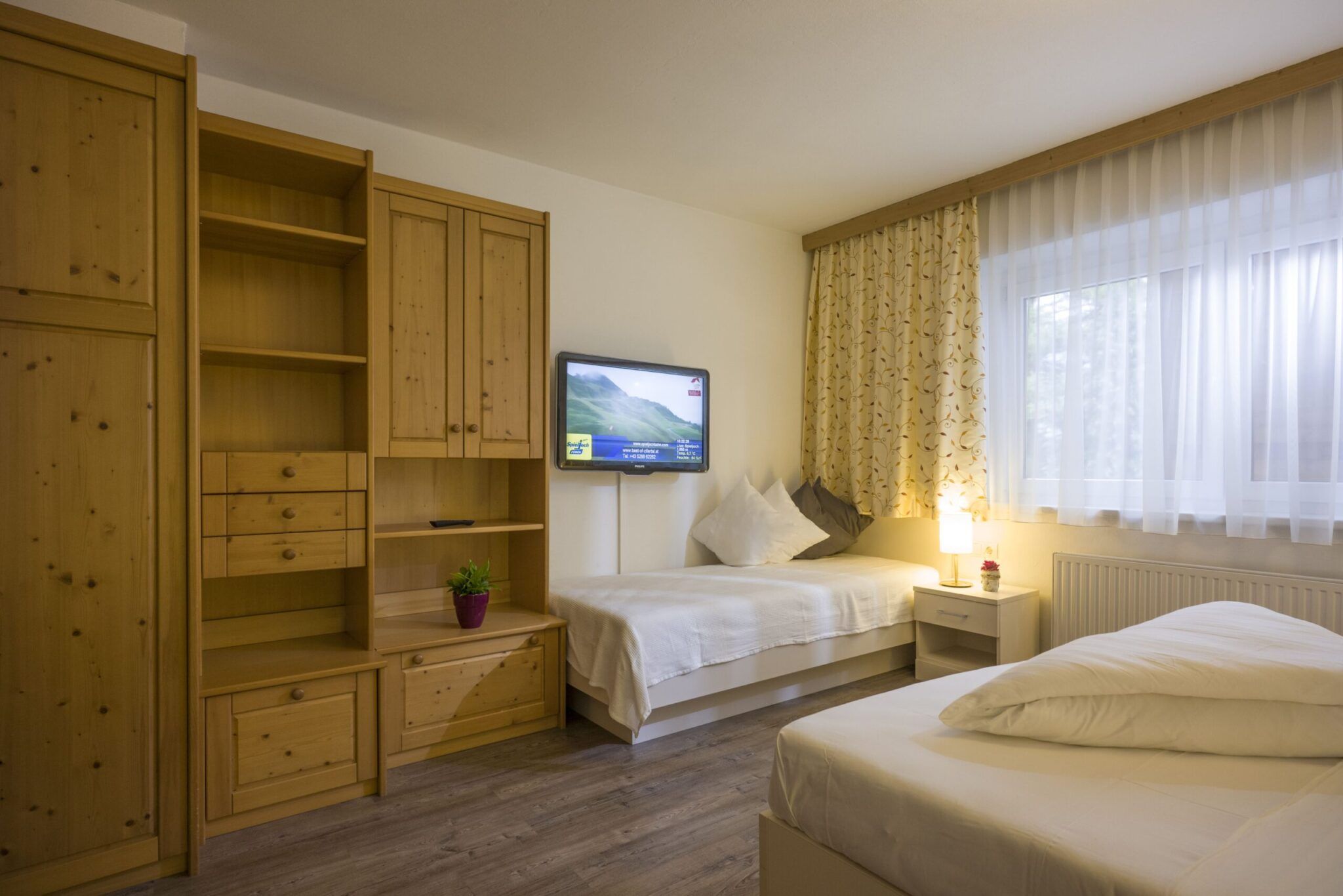 Ein gemütliches Schlafzimmer mit Holzmöbeln, einem Fernseher an der Wand, zwei Betten, einem Fenster mit Vorhängen und einem kleinen Nachttisch.