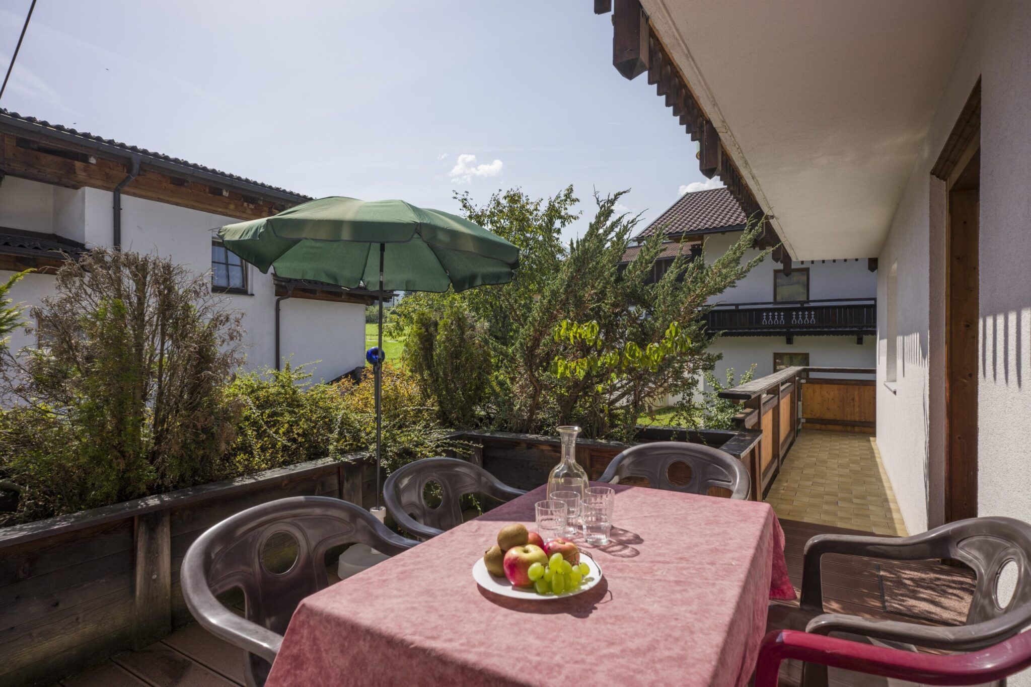 Ein sonnendurchfluteter Balkon mit einem Tisch mit Obst und Getränken, vier Stühlen, Topfpflanzen und einem grünen Sonnenschirm im Hintergrund.