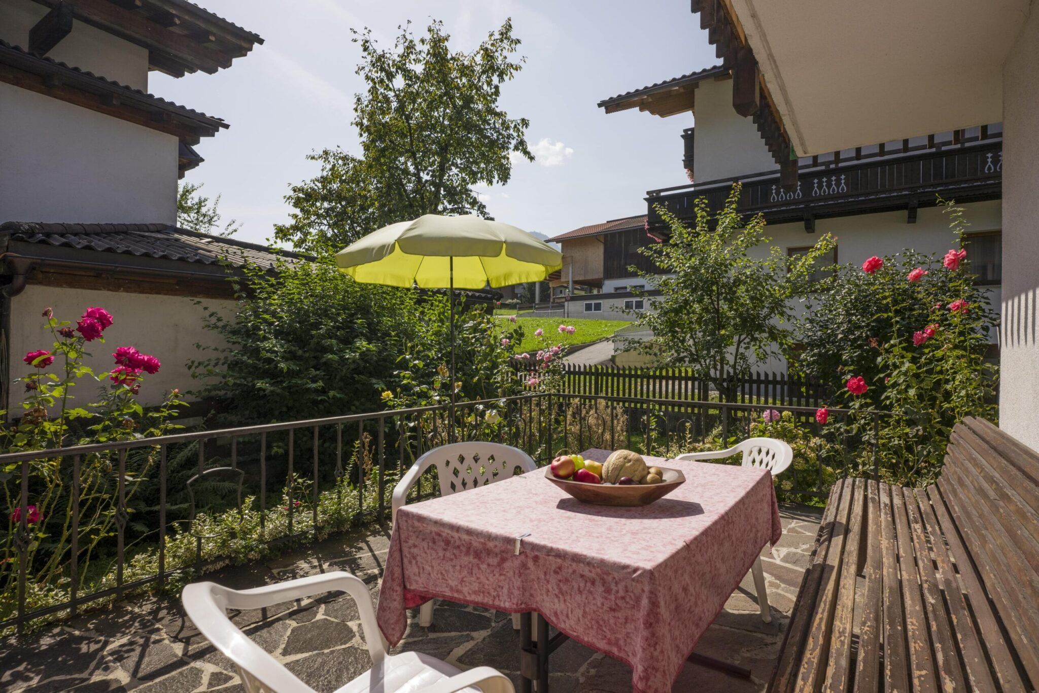Eine gemütliche Terrasse mit einem Tisch, bedeckt mit einer rosa Tischdecke, umgeben von Blumen und Grün und den benachbarten Holzhäusern.