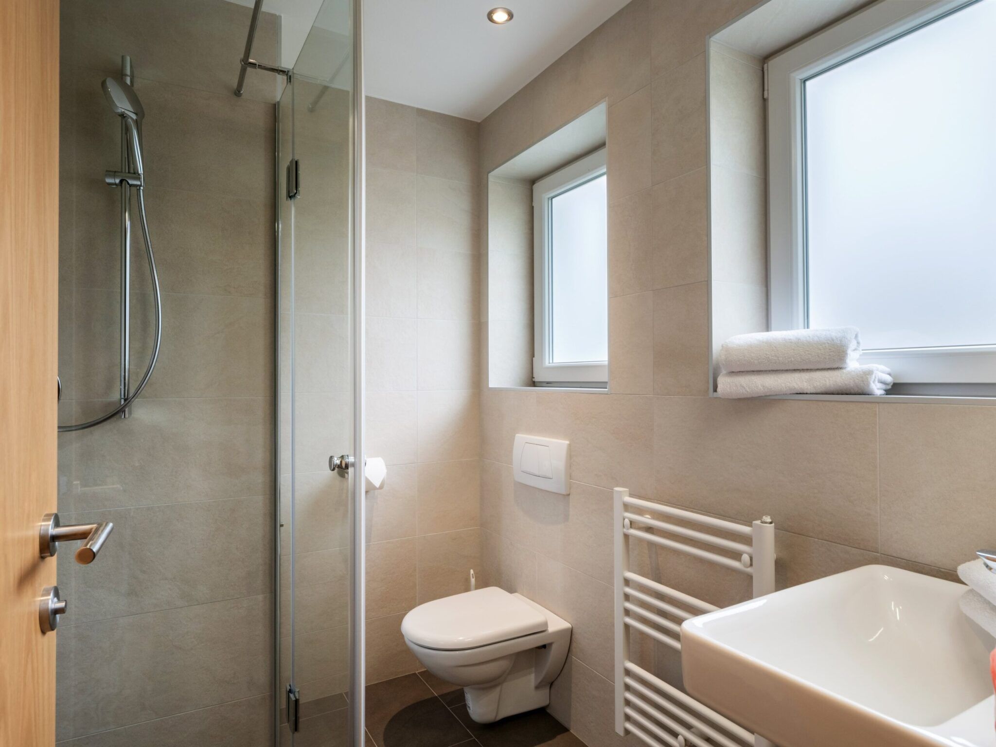 Modernes Badezimmer mit ebenerdiger Dusche, wandmontiertem WC, Handtuchhalter, Waschbecken und Milchglasfenstern.