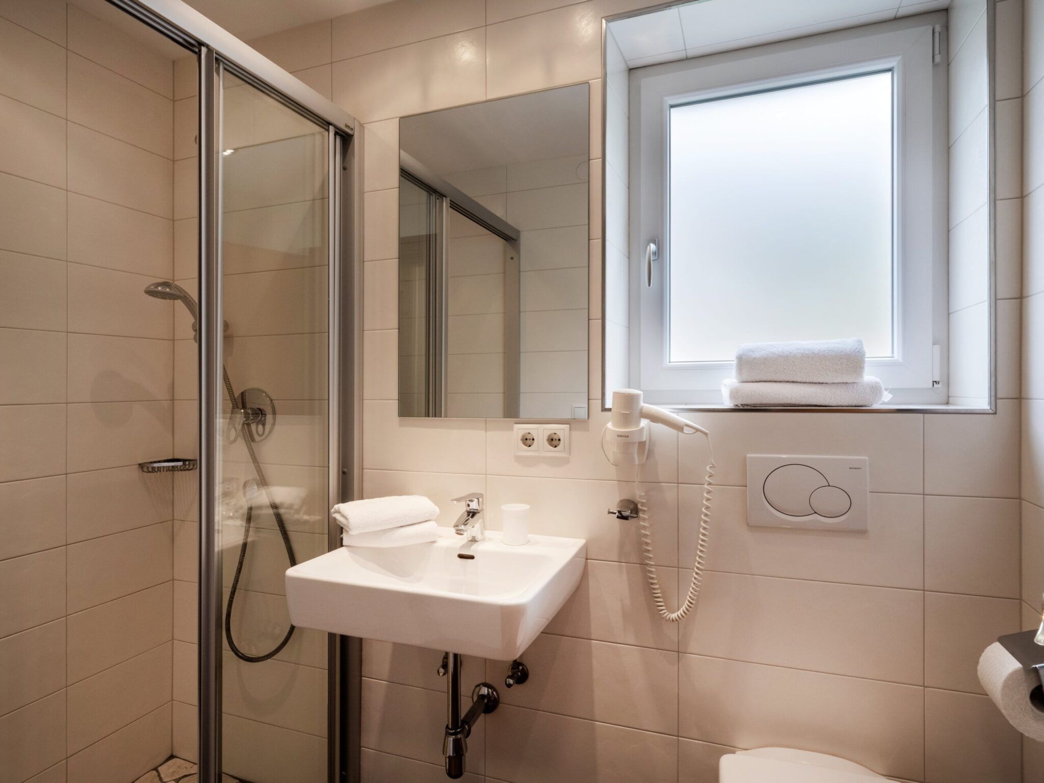 Modernes Badezimmer mit Glasdusche, weißem Waschbecken, großem Spiegel, wandmontiertem Haartrockner und Milchglasfenster.