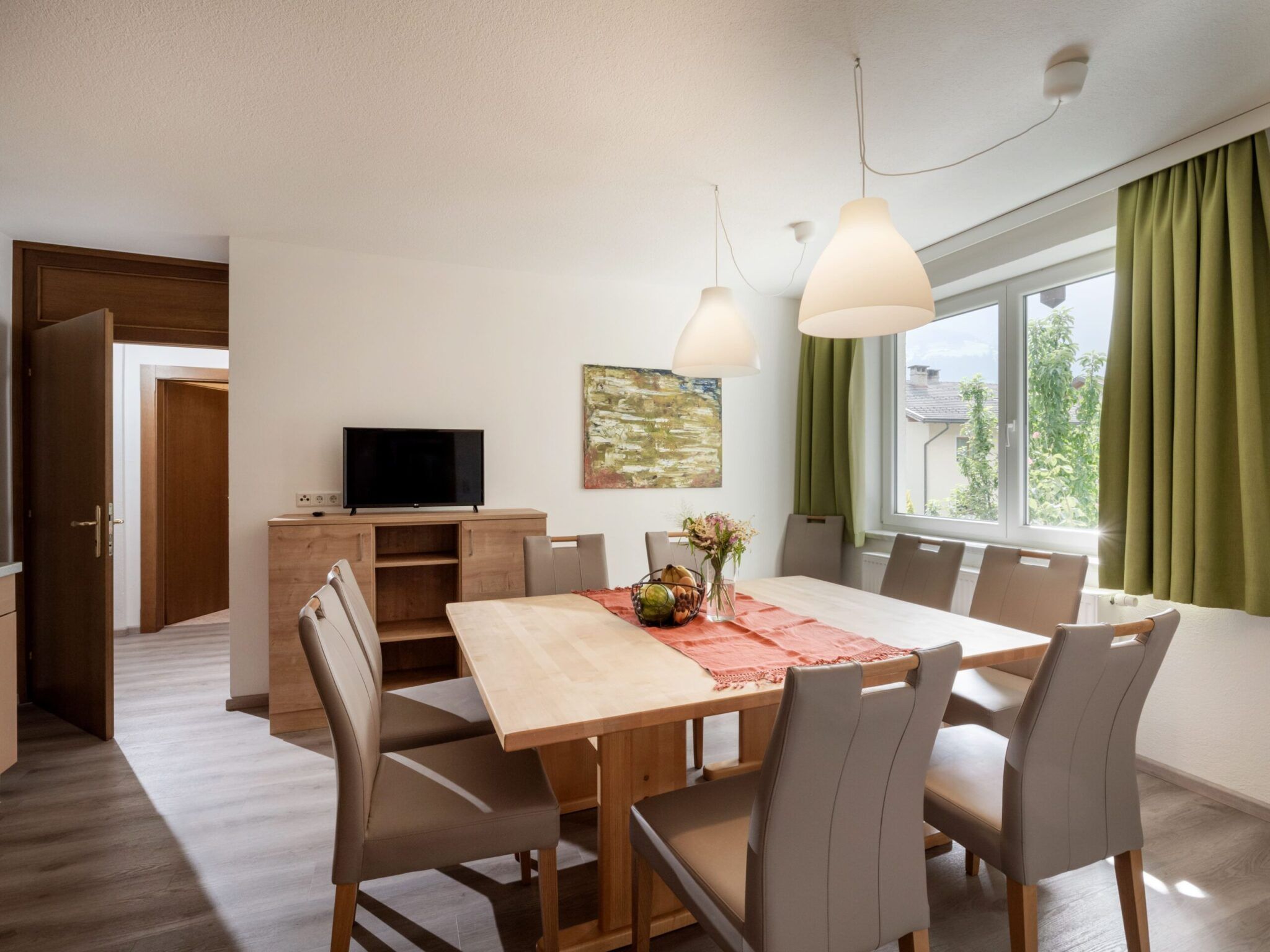 Esszimmer mit einem Holztisch, beigen Stühlen, Hängelampen, einem Fernseher auf einem Sideboard und einem großen Fenster mit grünen Vorhängen.