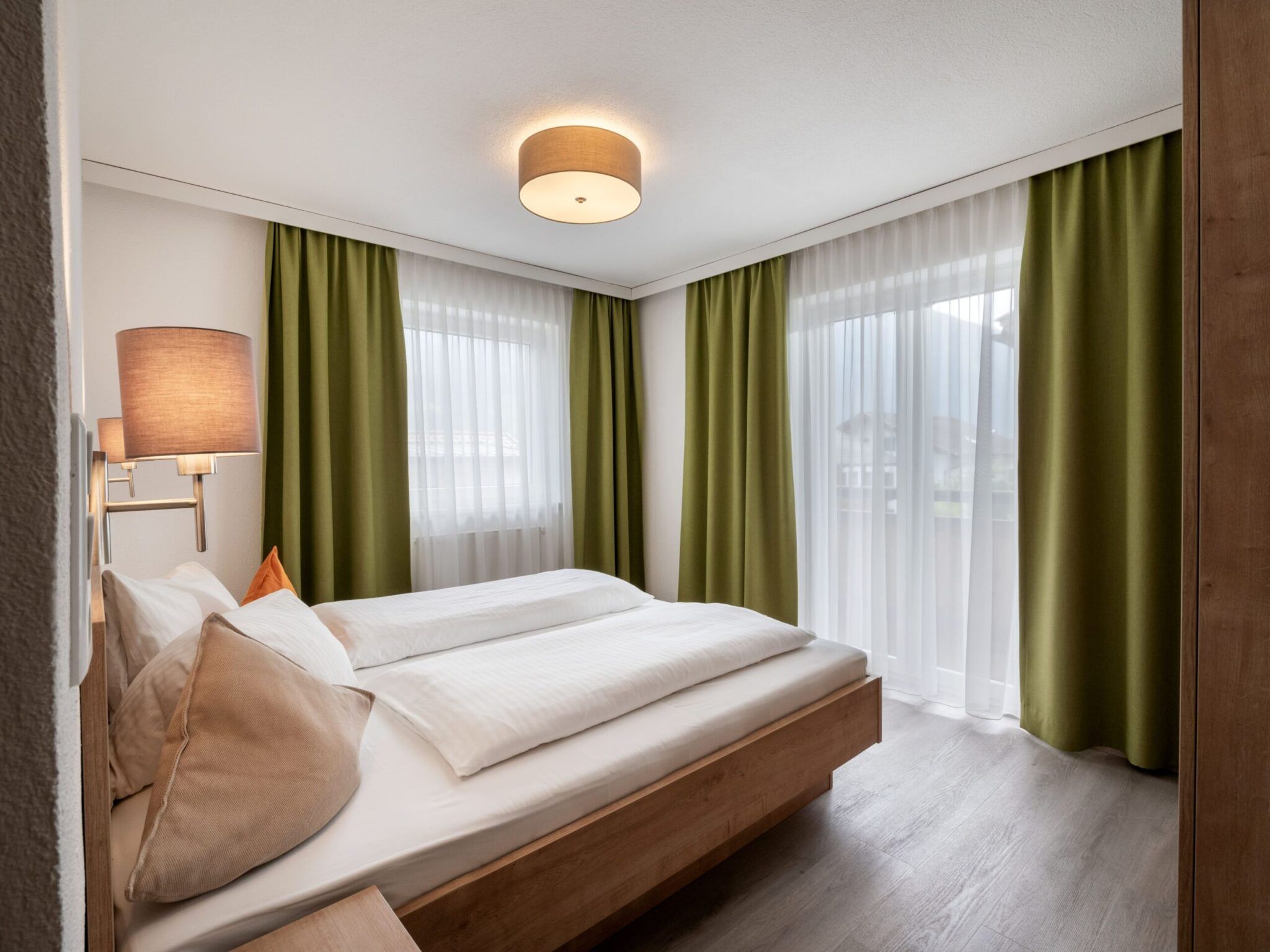 Ein gemütliches Schlafzimmer mit einem Holzbett, weißer Bettwäsche, grünen Vorhängen und einer Deckenleuchte.