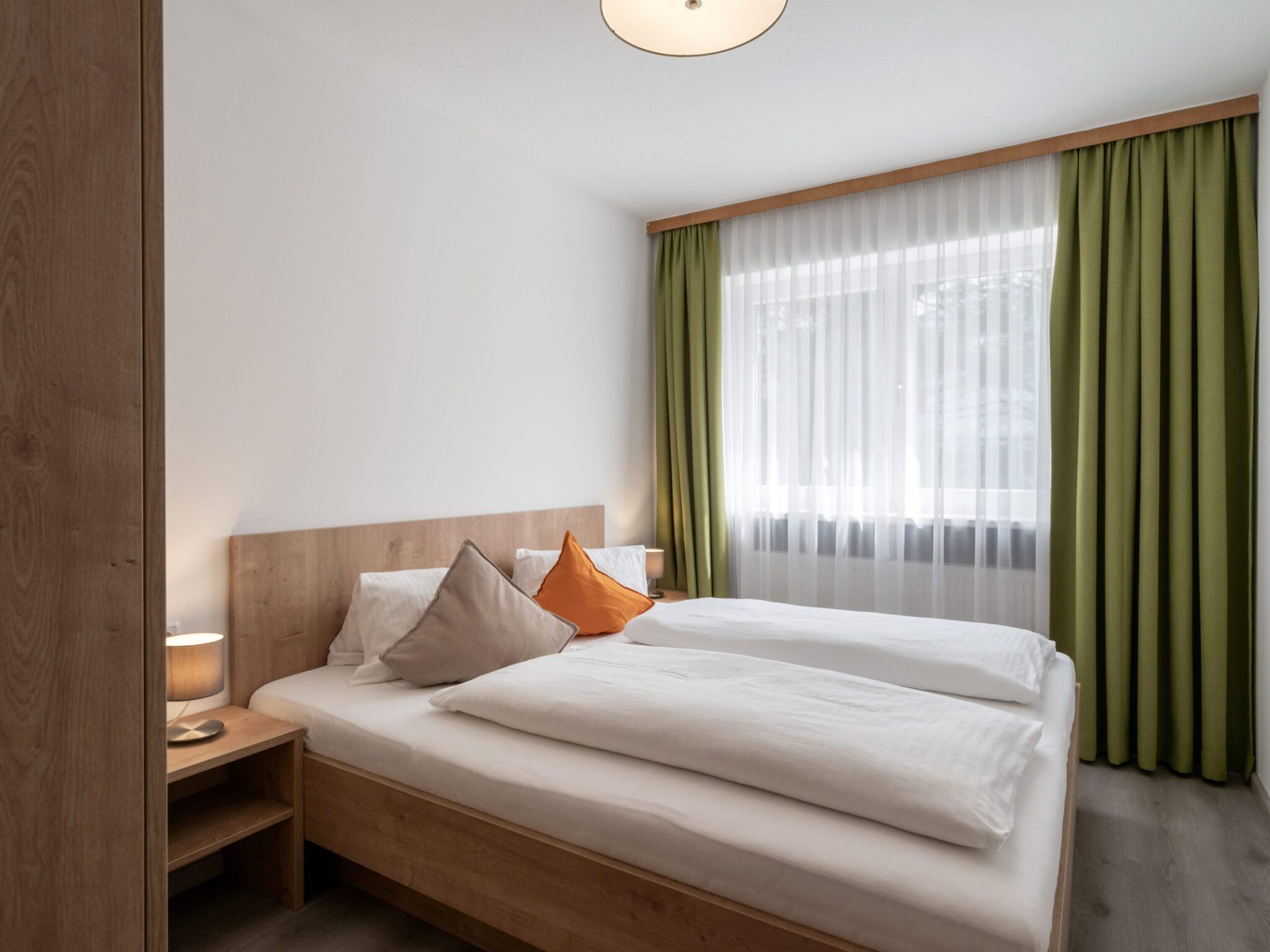 Ein minimalistisches Schlafzimmer mit zwei Betten, grünen Vorhängen und weißen Wänden. Links steht ein Nachttisch mit einer Lampe.