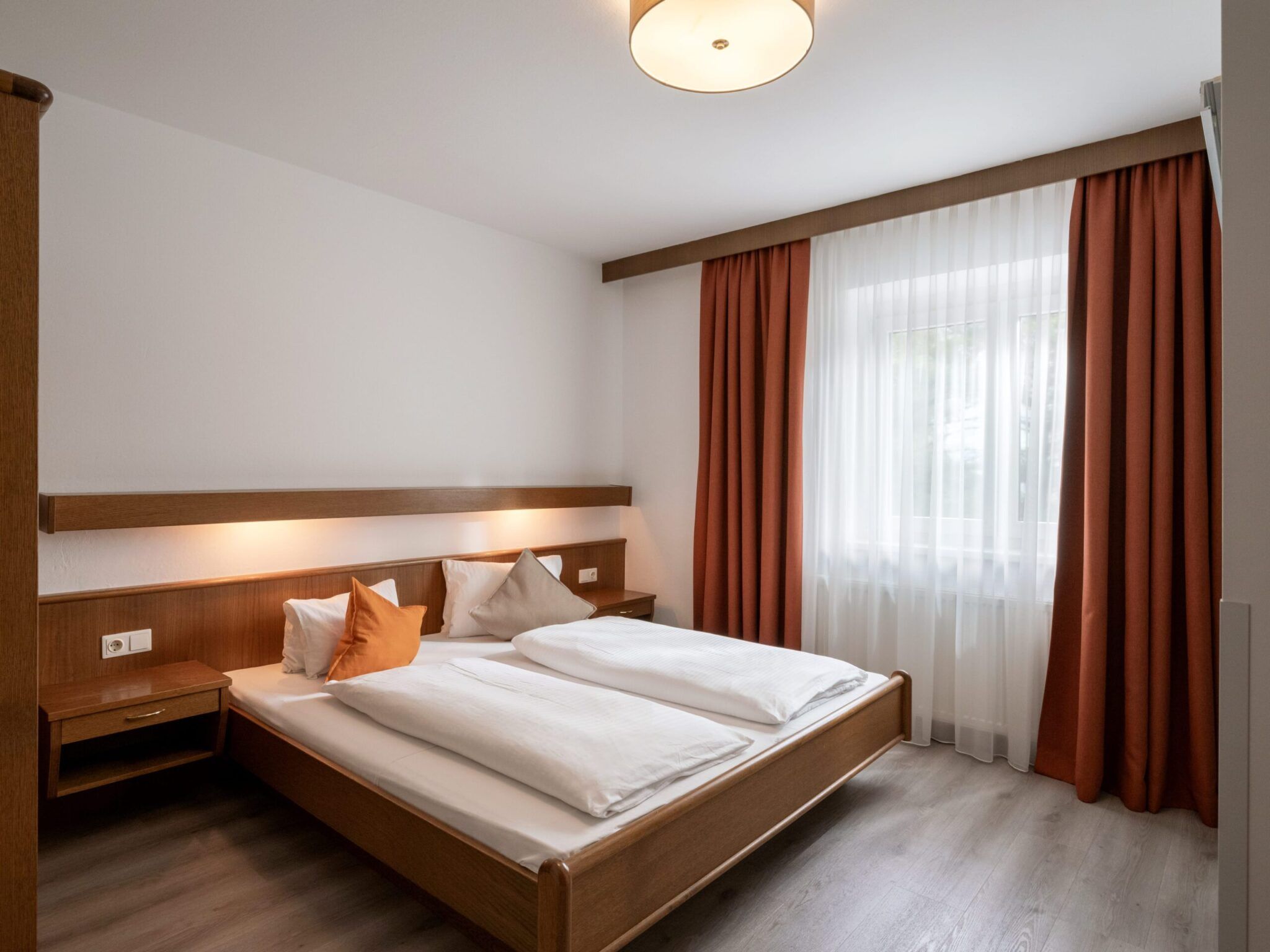 Ein ordentliches Schlafzimmer mit einem Doppelbett, weißer Bettwäsche, Holzmöbeln, roten Vorhängen und einem Fenster, das Tageslicht hereinlässt.