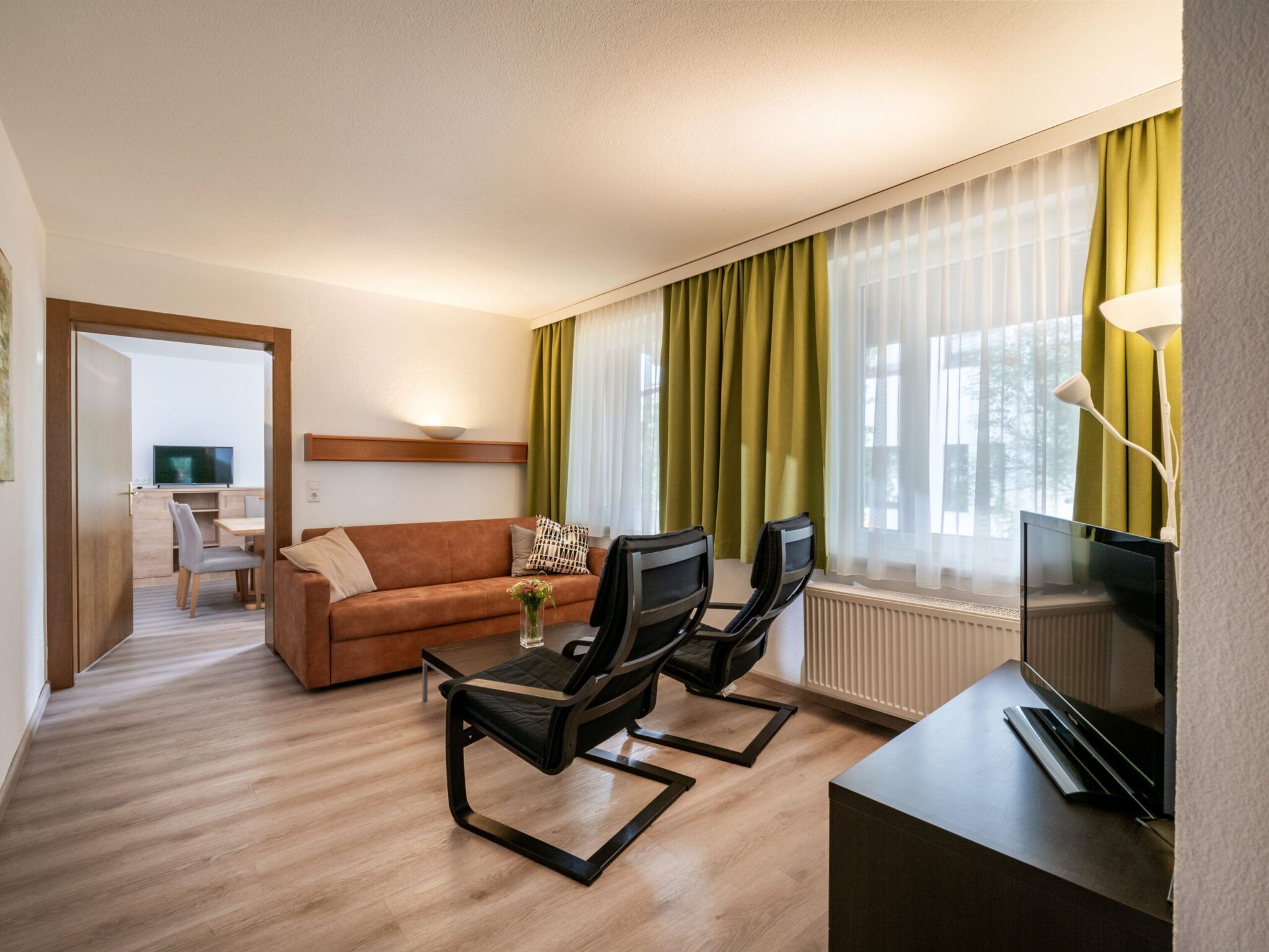 Modernes Wohnzimmer mit einem Sofa, zwei schwarzen Stühlen, einem Fernseher und einer Tür zu einem angrenzenden Raum. Grüne Vorhänge bedecken die Fenster.