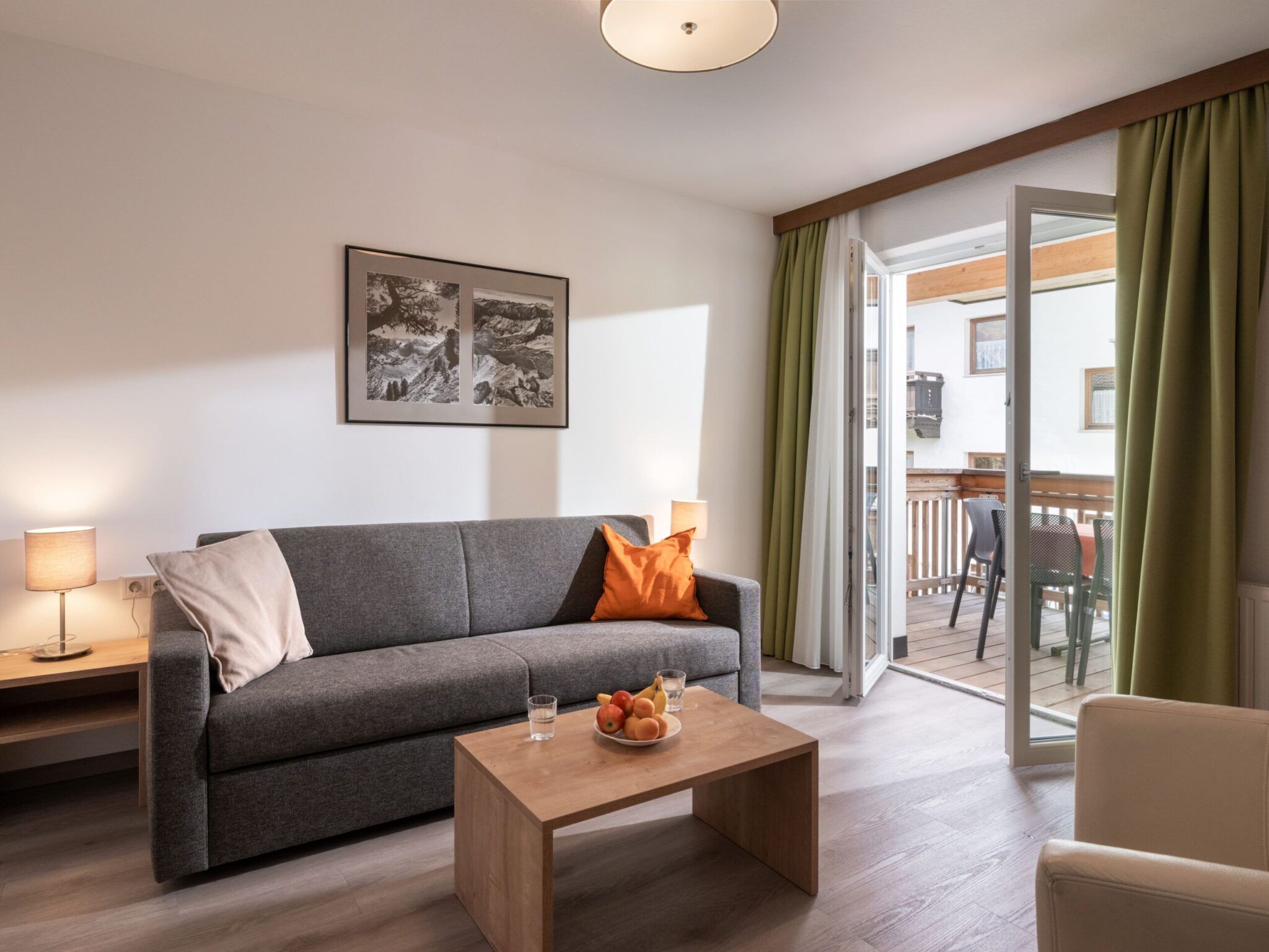 Ein gemütliches Wohnzimmer verfügt über ein graues Sofa, einen Couchtisch mit Obst, Zugang zum Balkon und minimalistische Wandkunst.