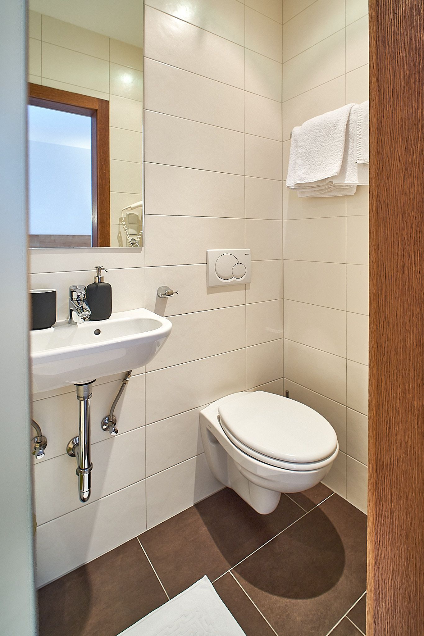 Modernes Badezimmer mit wandmontiertem Waschbecken, Toilette, Spiegel, Handtuchhalter mit gefalteten Handtüchern sowie gefliesten Wänden und Boden.