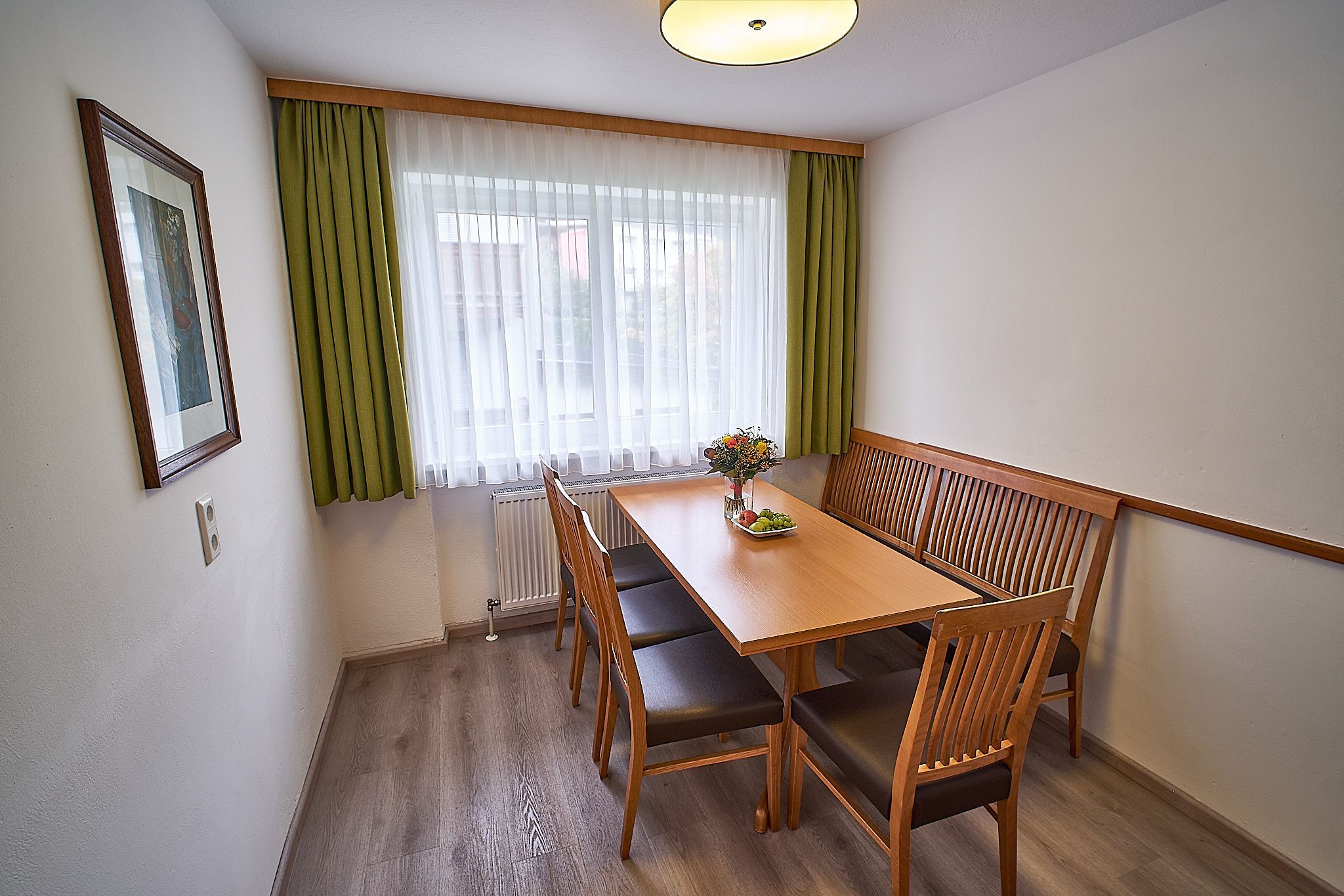 Gemütlicher Essbereich mit einem Holztisch, vier Stühlen, grünen Vorhängen und einer Blumenvase.