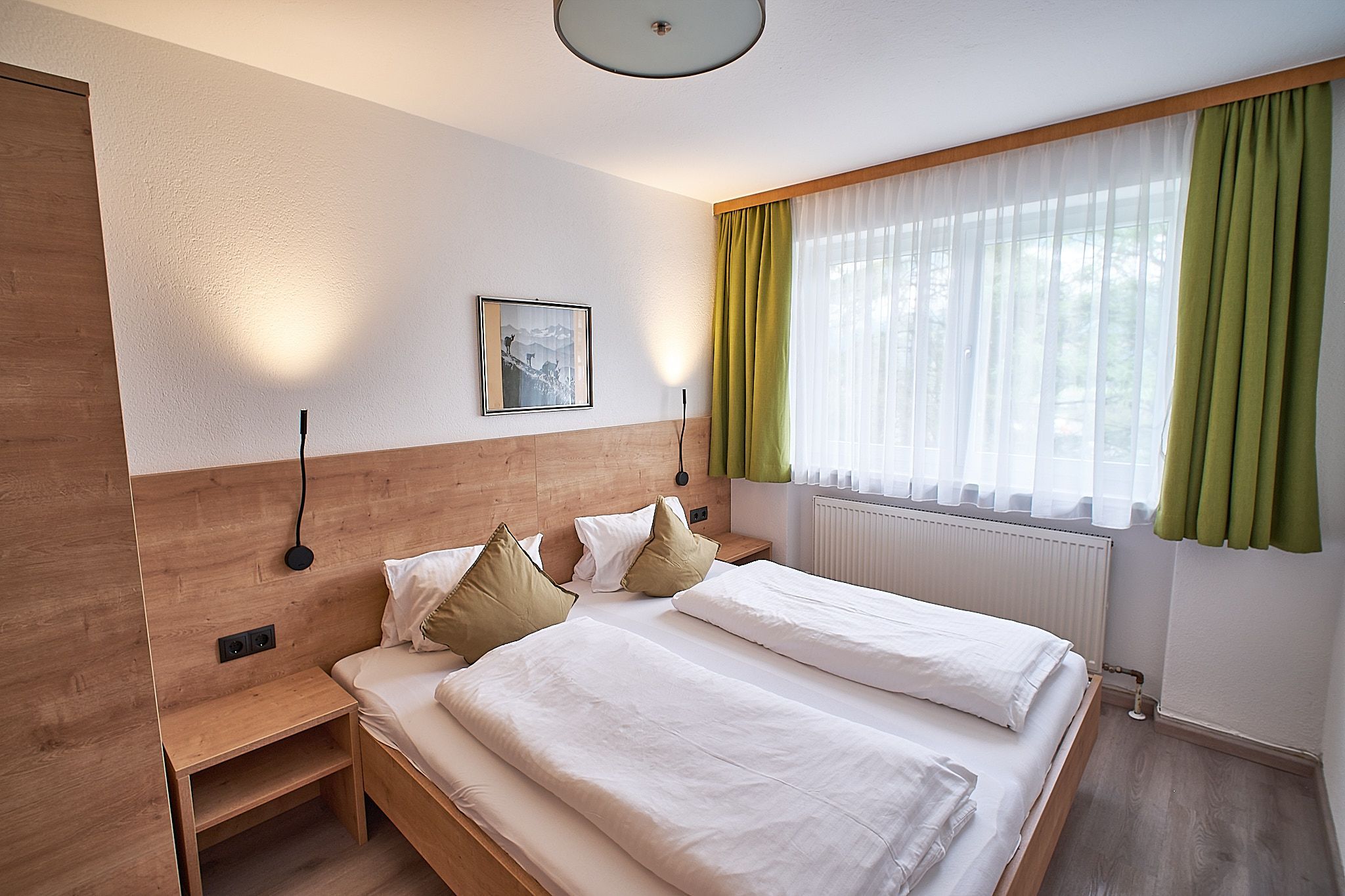 Gemütliches Schlafzimmer mit zwei Einzelbetten, Holzakzenten, grünen Vorhängen und einem großen Fenster, das Tageslicht hereinlässt.