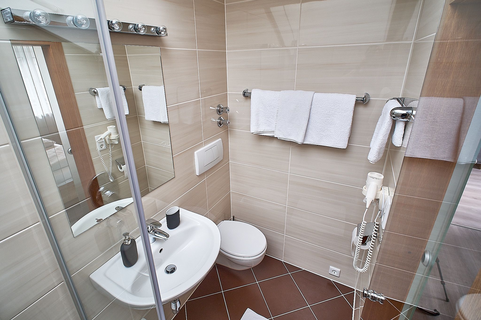 Ein kleines, modernes Badezimmer mit Waschbecken, Spiegel, Handtüchern und Haartrockner an beige gefliesten Wänden.