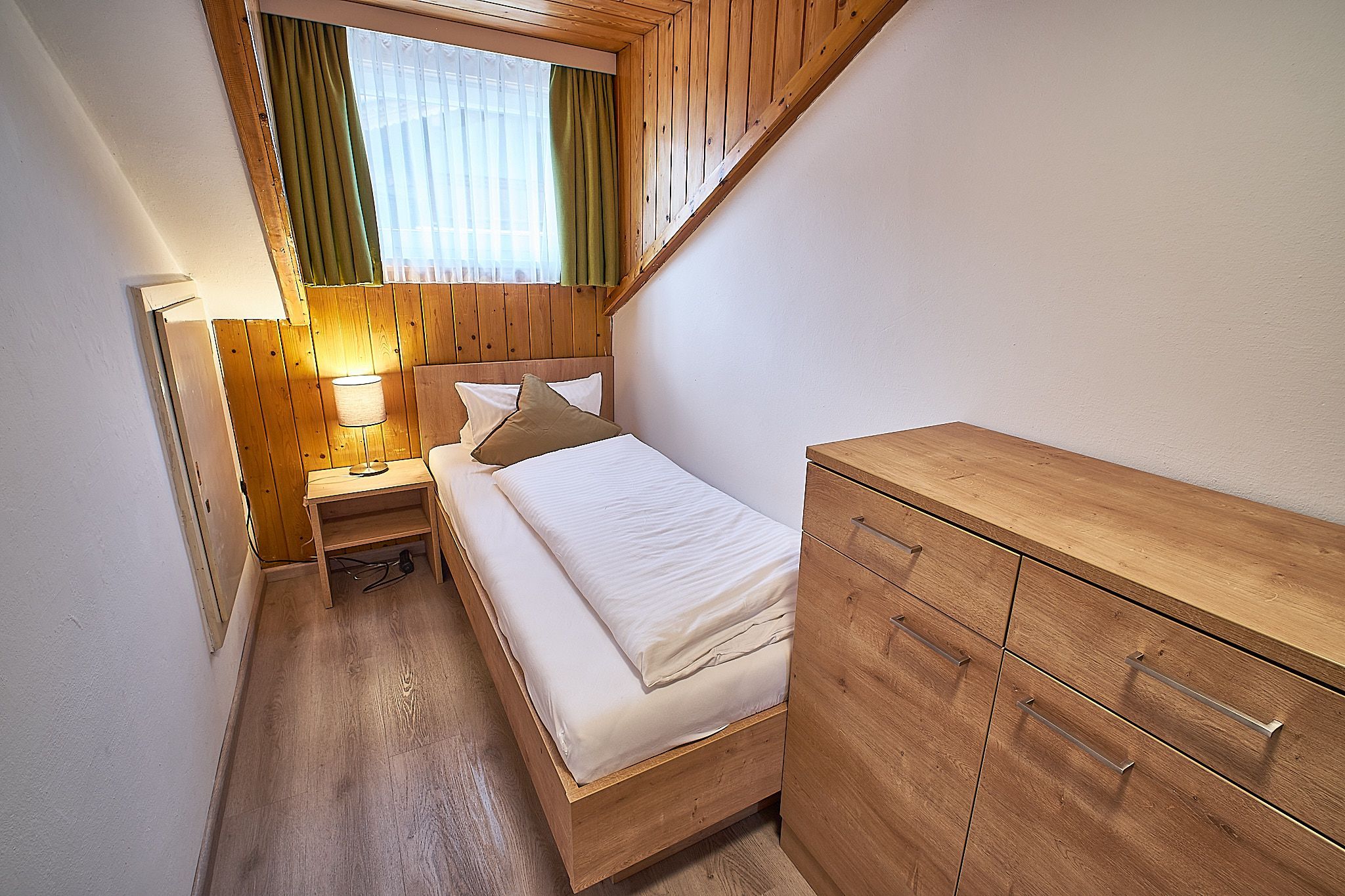Kleines Dachgeschossschlafzimmer aus Holz mit Einzelbett, Nachttisch, Lampe und Kommode.